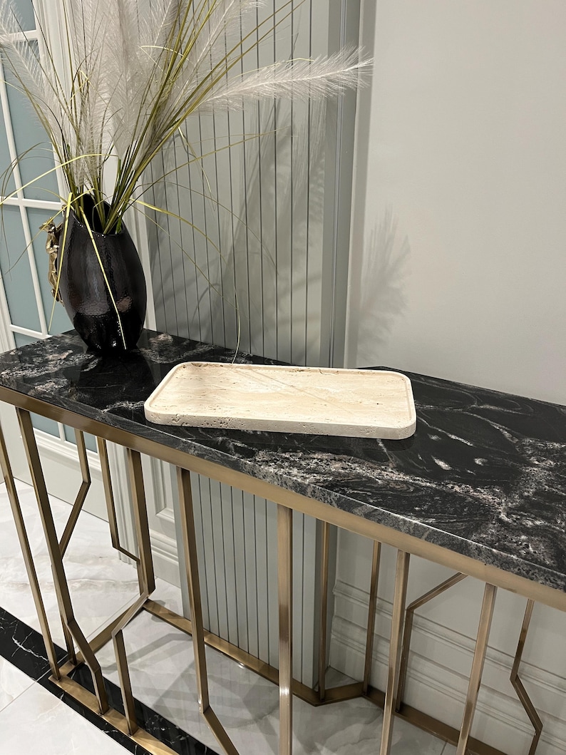 K&ouml;nnte beinhalten: Ein rechteckiges, hellbeiges Serviertablett aus Naturstein liegt auf einer schwarzen Granit-Arbeitsplatte mit goldenen Metallbeinen. Das Tablett befindet sich auf einem Konsolentisch mit geometrischem Design.