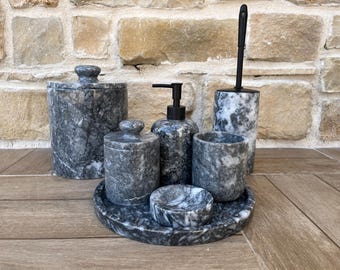 May include: A set of bathroom accessories made of gray marble. The set includes a soap dispenser, a toothbrush holder, a tray, a small bowl, two lidded jars, and a tumbler. The marble has a natural, veined pattern.
