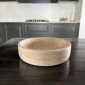 Puede incluir: Un cuenco redondo de piedra beige con una superficie texturizada. El cuenco est&aacute; sobre una mesa de madera marr&oacute;n oscuro. La cocina del fondo tiene armarios gris oscuro y una encimera blanca.