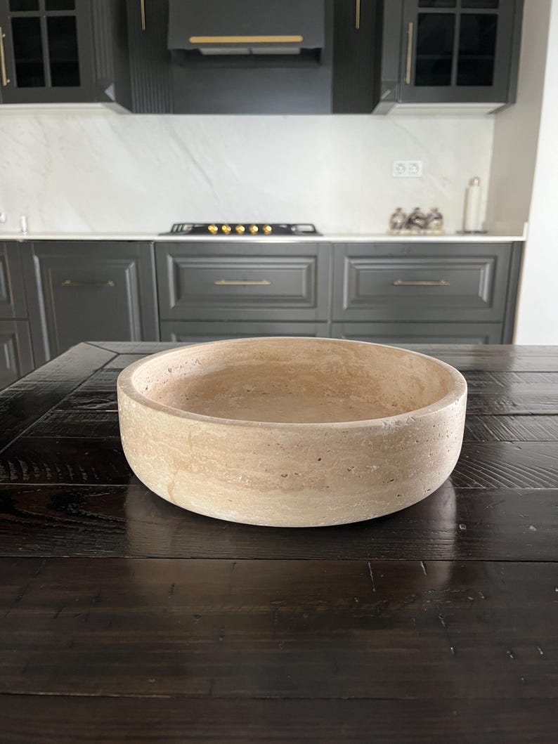 Puede incluir: Un cuenco redondo de piedra beige con una superficie texturizada. El cuenco est&aacute; sobre una mesa de madera marr&oacute;n oscuro. La cocina del fondo tiene armarios gris oscuro y una encimera blanca.