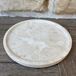 May include: A round, beige travertine coaster with a smooth, slightly textured surface. The coaster has a natural, organic look.