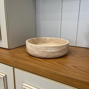 Puede incluir: Un cuenco redondo de piedra beige con una superficie texturizada. El cuenco est&aacute; sobre una superficie de madera, con un mueble en el fondo. El cuenco es un objeto decorativo.