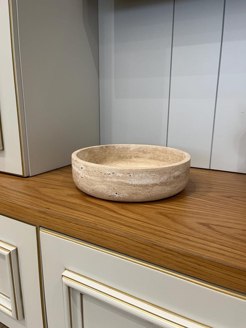 Puede incluir: Un cuenco redondo de piedra beige con una superficie texturizada. El cuenco est&aacute; sobre una superficie de madera, con un mueble en el fondo. El cuenco es un objeto decorativo.