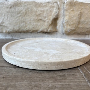 May include: A round, light beige, stone tray with a slightly raised edge. The tray is made of a natural material with a rough texture.