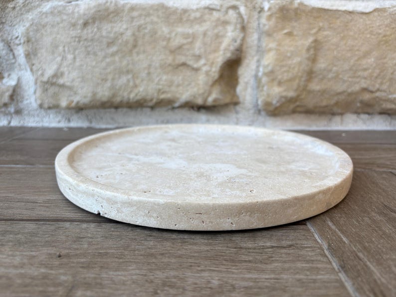 May include: A round, light beige, stone tray with a slightly raised edge. The tray is made of a natural material with a rough texture.