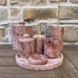 May include: A set of six bathroom accessories made of pink marble. The set includes a soap dispenser, toothbrush holder, tumbler, soap dish, a small jar with a lid, a larger jar with a lid, and a round tray.