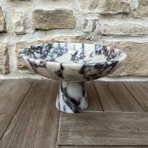 May include: A marble pedestal bowl with a white and purple veined pattern. The bowl has a wide, shallow dish shape and sits on a short, flared pedestal base. The bowl is displayed on a wooden surface in front of a stone wall.