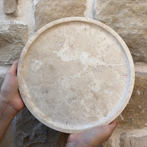 May include: A round, beige travertine serving tray with a slightly raised rim. The tray has a natural, textured surface.
