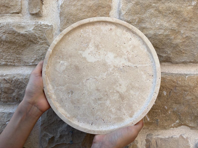 May include: A round, beige travertine serving tray with a slightly raised rim. The tray has a natural, textured surface.