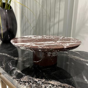 Puede incluir: Un soporte para tartas redondo de mármol con un diseño de mármol rojo y blanco. El soporte tiene una base de pedestal y está sentado sobre una superficie de mármol negro y blanco.
