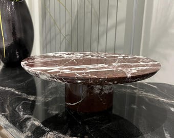 May include: A round marble cake stand with a red and white marbled design. The stand has a pedestal base and is sitting on a black and white marbled surface.