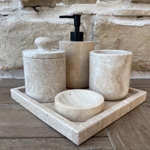 May include: A beige marble bathroom set comprising a soap dispenser, a tumbler, a lidded jar, a small dish, and a rectangular tray. The set is made of natural stone with a speckled pattern and a matte finish. The soap dispenser has a black pump.