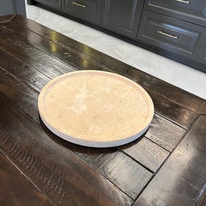 May include: A round, light brown, decorative tray made of natural stone sits on a dark wood table.