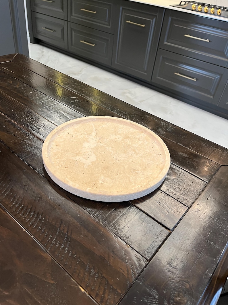 May include: A round, light brown, decorative tray made of natural stone sits on a dark wood table.