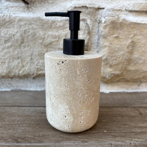 May include: A cylindrical soap dispenser with a beige, textured stone base and a black pump. The dispenser is set against a light-colored stone wall and a wooden surface. The dispenser is approximately 6 inches tall.