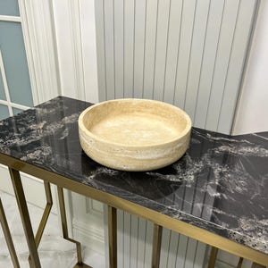 Puede incluir: Un cuenco redondo de piedra beige se encuentra sobre una mesa con vetas de m&aacute;rmol blanco y negro. La mesa tiene un marco de metal dorado. El cuenco es de color beige claro y tiene una superficie texturizada. La mesa est&aacute; frente a una pared blanca.