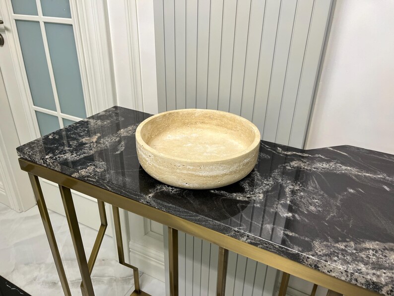 Puede incluir: Un cuenco redondo de piedra beige se encuentra sobre una mesa con vetas de m&aacute;rmol blanco y negro. La mesa tiene un marco de metal dorado. El cuenco es de color beige claro y tiene una superficie texturizada. La mesa est&aacute; frente a una pared blanca.