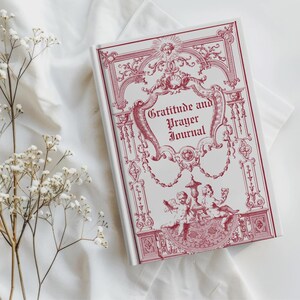 May include: A white journal with a red decorative design and the words "Gratitude and Prayer Journal" on the cover. The cover features ornate illustrations of cherubs, scrolls, and floral elements. The journal is set on a white cloth.