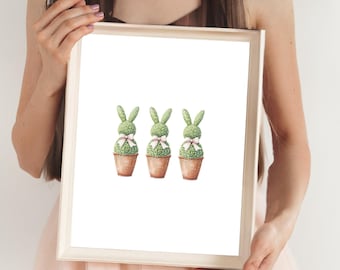 May include: A framed print featuring three topiary bunny sculptures in terracotta pots. Each bunny is green with a pink bow. The print has a white background and a light wood frame. The bottom right corner of the print has the text "@pinkskyprintables".