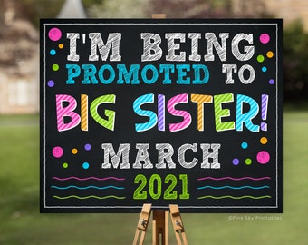 May include: A black chalkboard sign with colorful text announcing "I'm being promoted to big sister! March 2021". The sign is decorated with colorful dots and wavy lines.