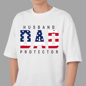 May include: White t-shirt with the words "HUSBAND DAD PROTECTOR" in black text. The word "DAD" is designed with a patriotic American flag pattern, featuring blue stars and red stripes.