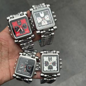 Könnte beinhalten: Vier quadratische Uhren mit silbernen Metallarmbändern. Die Zifferblätter der Uhren sind schwarz, rot und weiß, mit mehreren Zifferblättern und dem Markennamen "Oakley". Die Uhren werden vor einem grauen Hintergrund gehalten.