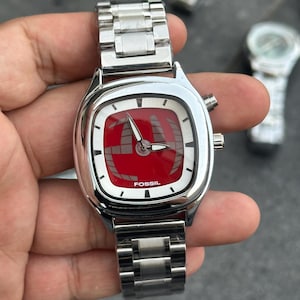 Peut inclure: Une montre-bracelet Fossil avec un bracelet en acier inoxydable et un cadran carré. Le cadran est rouge avec des détails blancs et des aiguilles noires. Le mot "FOSSIL" est imprimé sur le cadran.