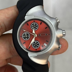 Könnte beinhalten: Eine silberne und schwarze Armbanduhr mit rotem Zifferblatt. Die Uhr hat drei Hilfszifferblätter und ein schwarzes Kautschukarmband. Die Uhr wird in einer Hand gehalten.