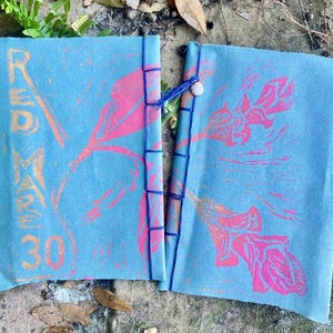 May include: Two open, light blue fabric journals with pink floral designs and blue stitching. The left journal has the text "RED MARE 30" in orange. A small seashell adorns the binding. The journals are lying on a natural surface.