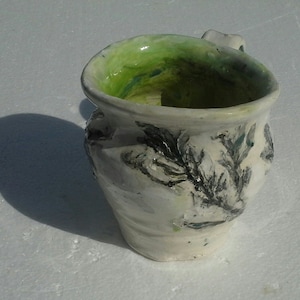 May include: A white ceramic mug with a green glaze inside and black and green floral designs on the outside.