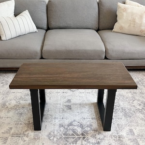 May include: A rectangular coffee table with a dark brown wooden top and black metal legs. The table is positioned in front of a gray sofa with decorative pillows. The rug beneath the table has a gray and white pattern.