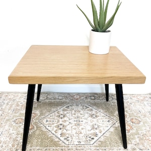 May include: A square wooden end table with a light wood grain finish and black tapered legs. The table is sitting on a patterned rug and has a small potted plant on top.