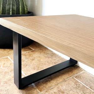 May include: A light-colored wooden table with a black metal frame. The tabletop has a wood grain pattern. The table is supported by a black, angular metal base. The table is placed on a tiled floor.