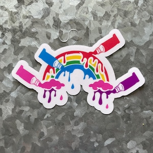 May include: A colorful sticker featuring a rainbow with paint dripping from paint tubes in red, blue, pink, and purple. The sticker has a white border and a clear plastic loop at the top for hanging.