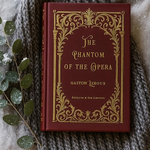 May include: A burgundy hardback book titled "The Phantom of the Opera" by Gaston Leroux, with gold decorative lettering and detailing. The book rests on a grey knitted blanket, with a sprig of greenery to the side.