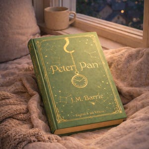 May include: A green hardback book titled "Peter Pan" by J.M. Barrie, with gold lettering and illustrations, lies on a soft blanket near a window. A mug is placed close by. The book's cover features a clock and constellation designs.