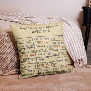 May include: A cushion with a vintage library card design. The cushion features the text "English & Ink Library DATE DUE" and a list of dates. The cushion is beige with black text and is placed on a bed.