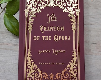 May include: A hardcover book titled The Phantom of the Opera by Gaston Leroux, English & Ink Edition. The cover is deep maroon with gold foil filigree borders and lettering. The title and author name are centered in gold text.