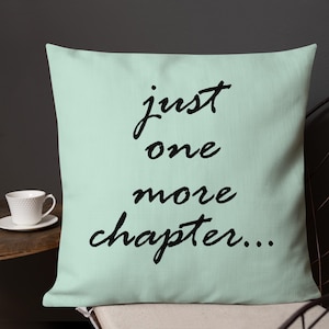 Op de afbeelding: Een vierkant, lichtgroen kussen met de tekst "just one more chapter..." in zwart script. Het kussen ligt op een stoel, met een witte theekop en schotel zichtbaar op de achtergrond.
