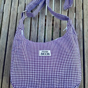 May include: A purple and white gingham shoulder bag with a long strap. The bag has a rounded shape and is made of fabric. The text "ATELIER 2MOON" is printed on a white label on the front of the bag.