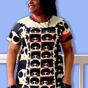 May include: A short-sleeved t-shirt with a repeating pattern of black-haired figures wearing sunglasses on a cream and black checkered background. The shirt has a red neckline and black sleeves. The person is wearing black gloves.