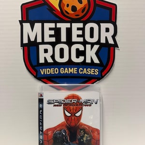 Pode incluir: Uma caixa de jogo de vídeo para PlayStation 3 de Spider-Man: Web of Shadows com uma máscara de Spider-Man vermelha e azul, Venom e Wolverine na capa. Acima da caixa está um logótipo da Meteor Rock Video Game Cases com um meteoro laranja.