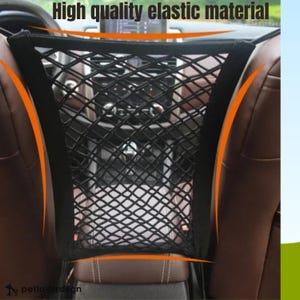 May include: Black mesh car storage net installed between the front seats. The text "High quality elastic material" is visible at the top. The net is designed to hold items and keep them secure while driving. Brown leather car seats are visible on either side.