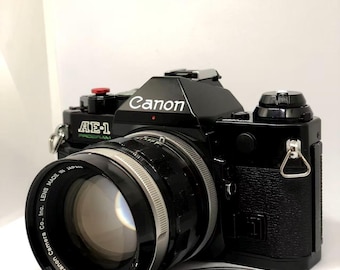 Pode incluir: Câmera Canon AE-1 Program preta com lente e tampa da lente. A câmera tem o logotipo da Canon em branco e o texto "AE-1 PROGRAM" em branco. A lente é preta e prateada, com o texto "Canon Camera Co., Inc. LENS MADE IN JAPAN".