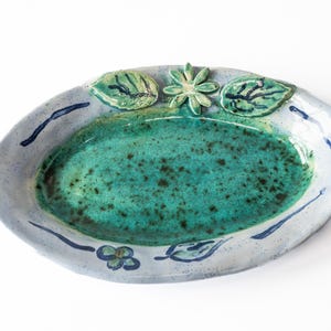 Può includere: Piatto da portata ovale in ceramica con interno verde acqua macchiato e bordo azzurro. Il bordo è decorato con pennellate blu scuro e accenti di foglie e fiori verdi. Un pezzo decorativo per la cucina o la sala da pranzo.