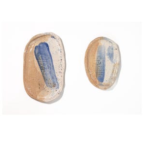 Könnte beinhalten: Zwei handgefertigte Keramikschalen mit organischen Formen. Die Schalen sind beige mit blauen Pinselstrichen und gesprenkelten Details. Das Design ist minimalistisch und künstlerisch, zum Servieren oder Ausstellen.