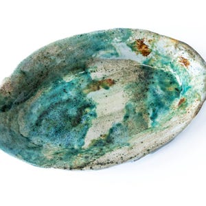 Op de afbeelding: Een ovaalvormige keramische schaal met een getextureerd oppervlak. De schaal heeft een mix van turquoise, bruine en witte kleuren. De binnenkant heeft een glanzende afwerking, terwijl de rand een ruwe, ongeglazuurde uitstraling heeft. Dit handgemaakte aardewerk is ideaal om te serveren of tentoon te stellen.