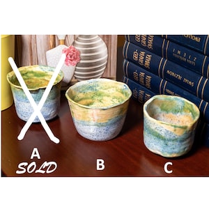 Könnte beinhalten: Drei handgefertigte Keramikbecher mit einer gesprenkelten blauen, gelben und grünen Glasur. Der Becher links ist mit "SOLD" und einem großen weißen "X" markiert. Die anderen beiden Becher werden auf einer dunklen Holzoberfläche ausgestellt.