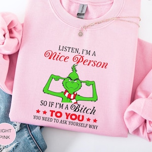 Op de afbeelding: Lichtroze sweatshirt met de tekst "LISTEN, I'M A Nice Person" in rode letters. Daaronder staat een groen stripfiguur met een rood-witte sjaal. De tekst "SO IF I'M A Bitch TO YOU YOU NEED TO ASK YOURSELF WHY" staat ook op het shirt.