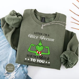 Op de afbeelding: Een olijfgroene sweatshirt met de tekst "LISTEN, I'M A Nice Person" en "SO IF I'M A Bitch TO YOU YOU NEED TO ASK YOURSELF WHY". De afbeelding toont een groen stripfiguur met een rood-witte sjaal.